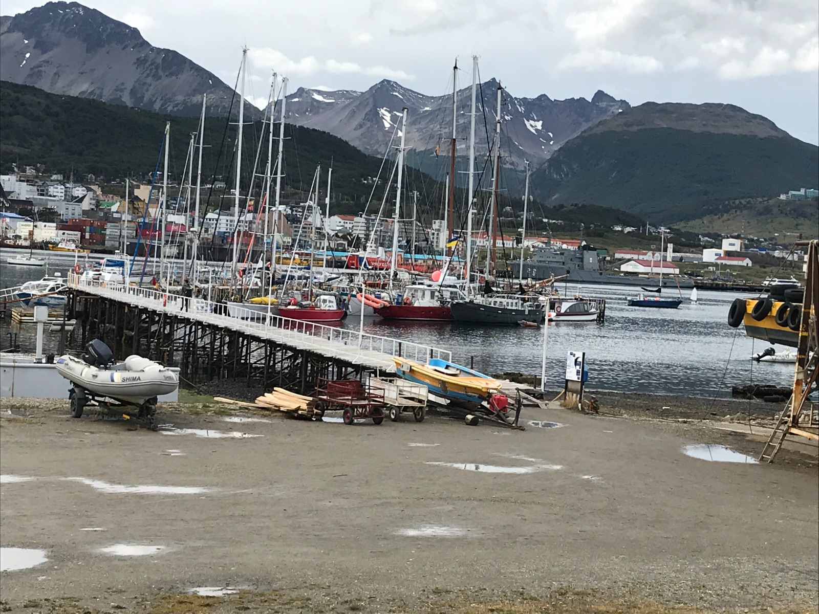 The departure port. Expedition yachts rafted along a trestle jetty, dinghies and timber ashore, a naval vessel on the far side.