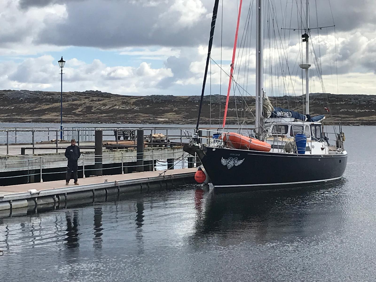 Mon Coeur alongside at Stanley — dark hull, winged-heart device on the bow with her name above it, RIB and water drums stowed on deck. The ship names on the hillside behind are Stanley's.