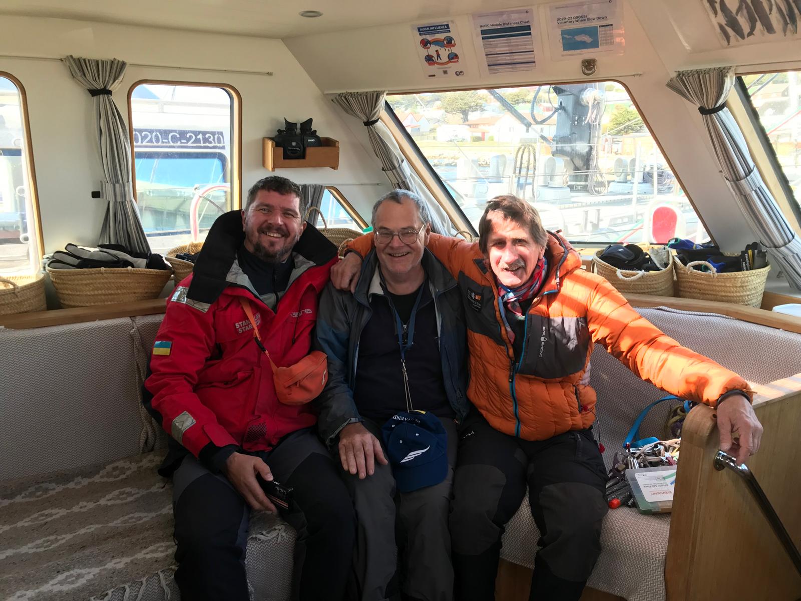 Three of the crew in a wheelhouse. Pinned above them: an avian influenza notice, an IAATO wildlife distances chart, and a GSGSSI voluntary whale slow-down notice headed 2022-23.