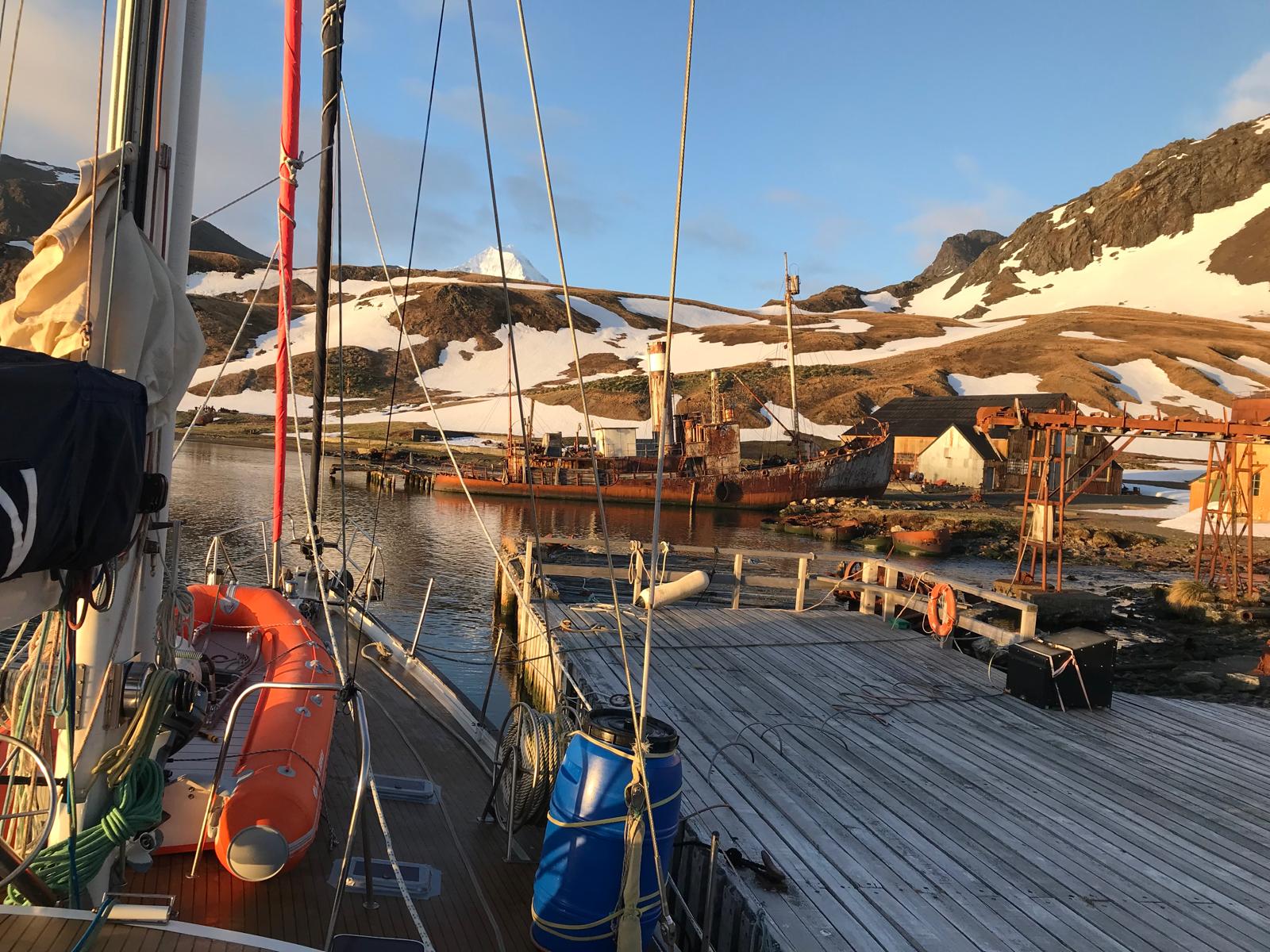 A beached whale catcher seen over the bow, her funnel and mast still standing. Behind, the station sheds and a hanging snowfield.